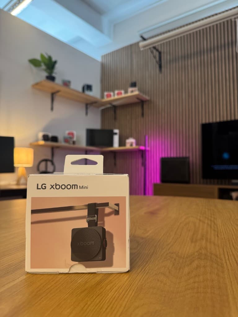 LG XBOOM Mini packaging on the circular oak table, with the slat wall and open shelving sets behind.